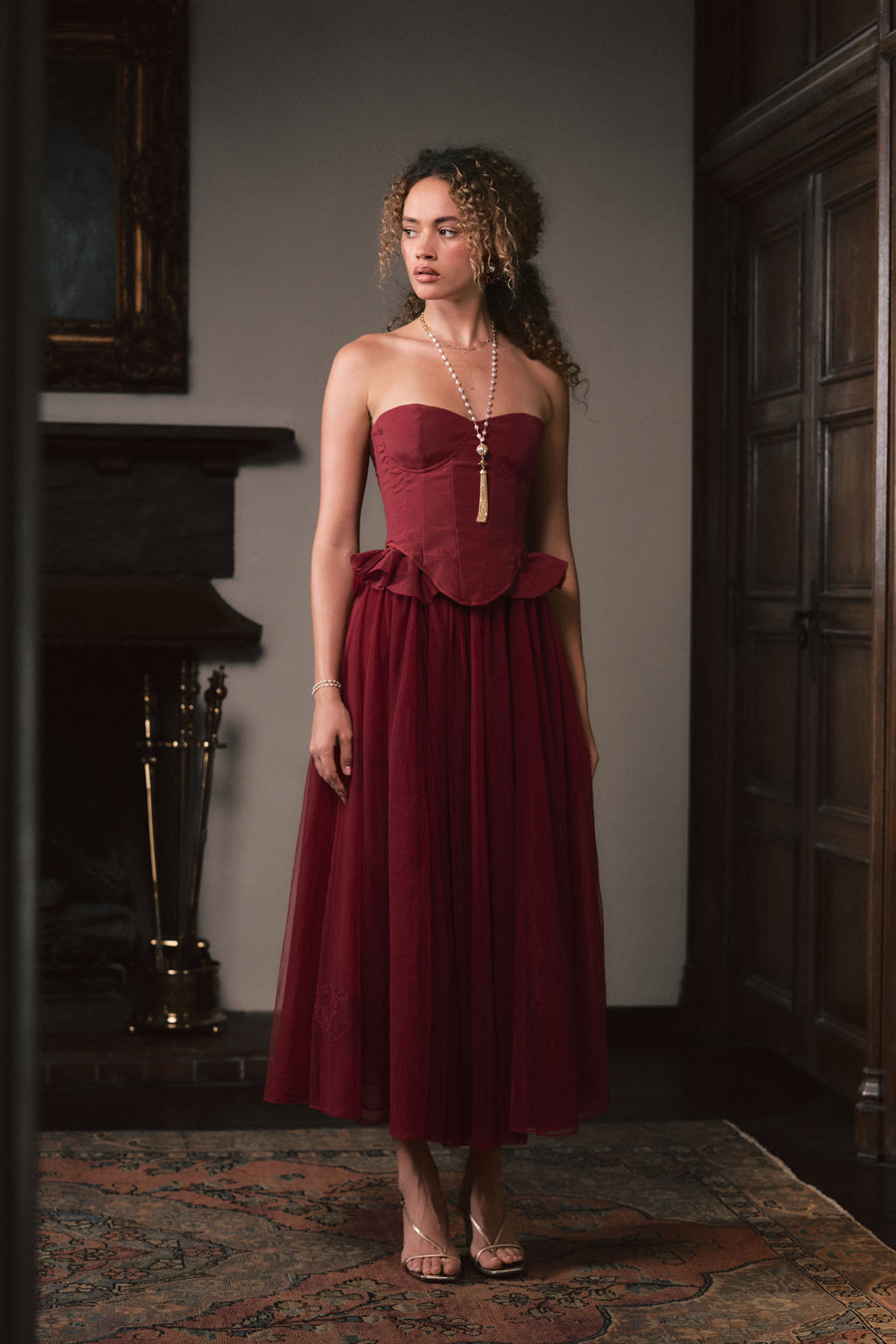 A woman stands indoors wearing a strapless burgundy midi dress with a structured bodice, ruffled waist, and flowing skirt. The room has dark wood paneling, a fireplace, and a patterned rug.