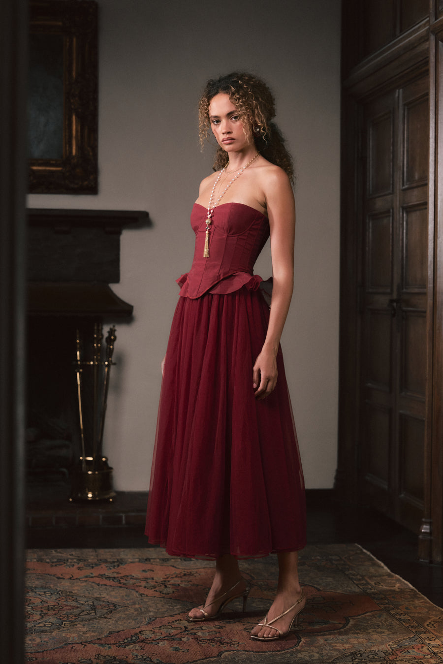 A woman stands indoors wearing a strapless burgundy midi dress with a structured corset-style bodice, peplum waist, and full gathered skirt. The room has dark wood paneling, a fireplace, and a patterned rug.