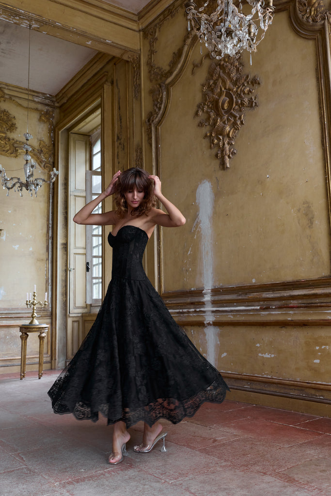 Woman in a black corset top and skirt standing in an ornate room with gold walls and chandeliers.