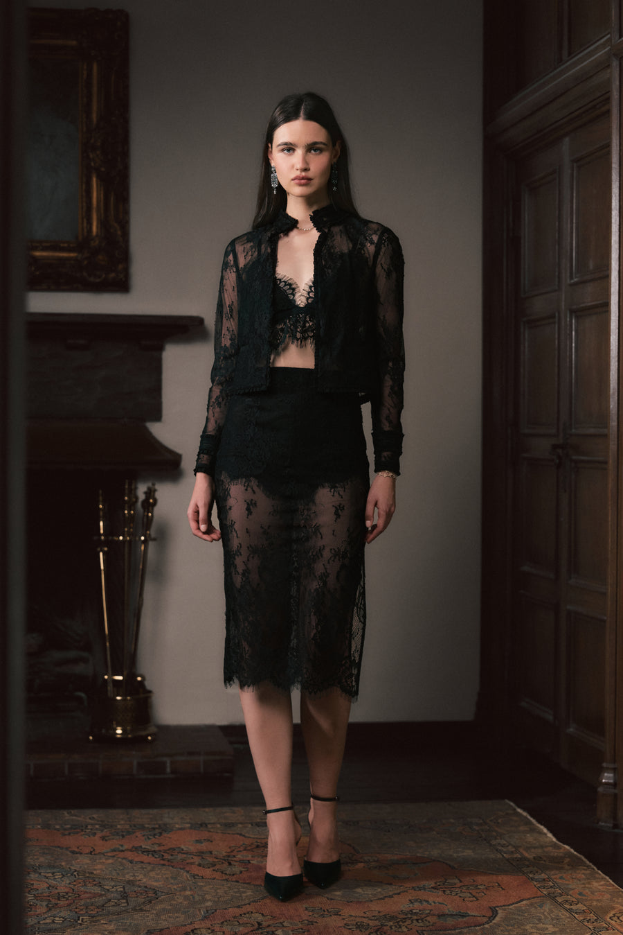 A woman stands in a dimly lit interior wearing a black sheer lace outfit consisting of a cropped long-sleeve top, a matching bralette, and a midi skirt, paired with black heels.