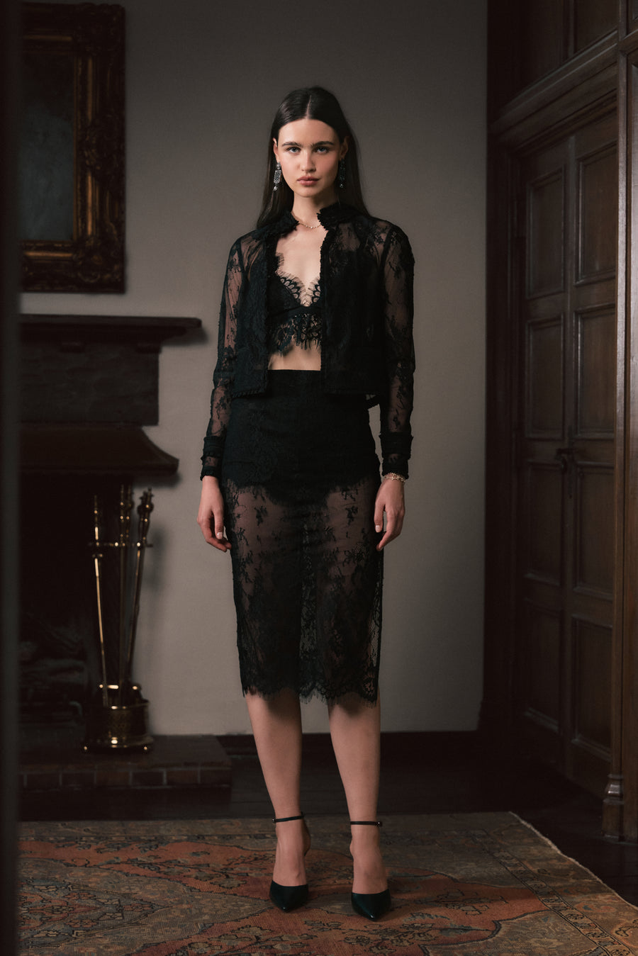 A model stands in a dimly lit room wearing a sheer black lace three-piece outfit consisting of a cropped jacket, a bralette, and a knee-length skirt, paired with black heels.