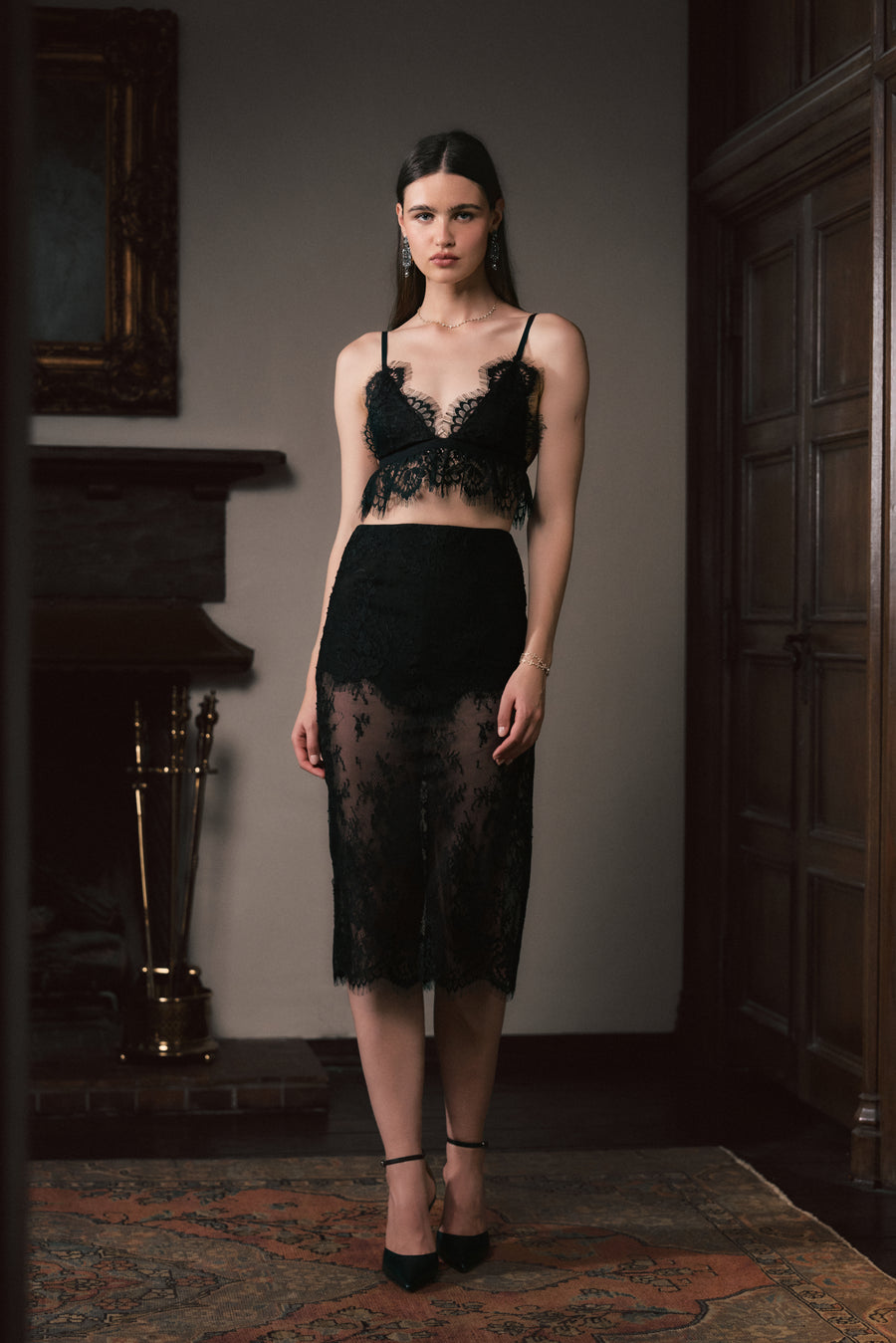A model stands in a dimly lit room wearing a black lace lingerie set consisting of a sheer underwire bra and a matching sheer midi skirt.