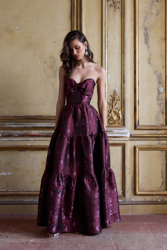 Woman in a burgundy evening gown standing in an ornate room with gold paneling.