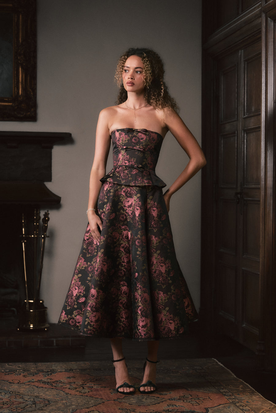 A woman stands indoors wearing a strapless midi dress with a fitted corset-style bodice and full skirt. The dress has a dark base with a pink floral jacquard pattern.