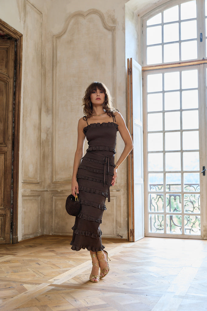 Woman in a dark brown dress standing in a room with large windows.