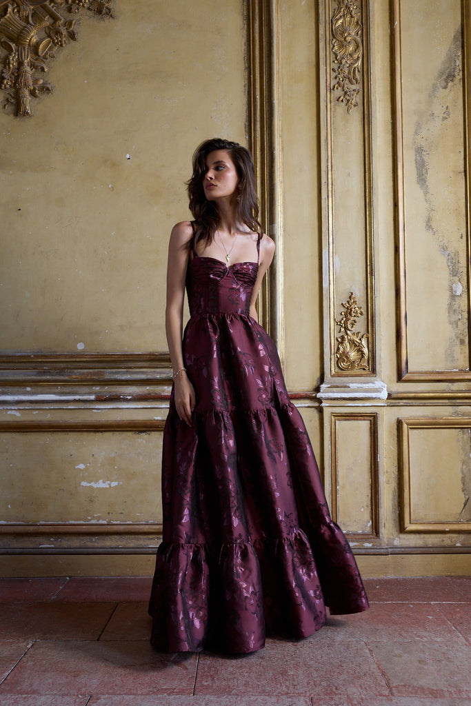 Woman in a burgundy evening gown standing in an ornate room with gold accents.