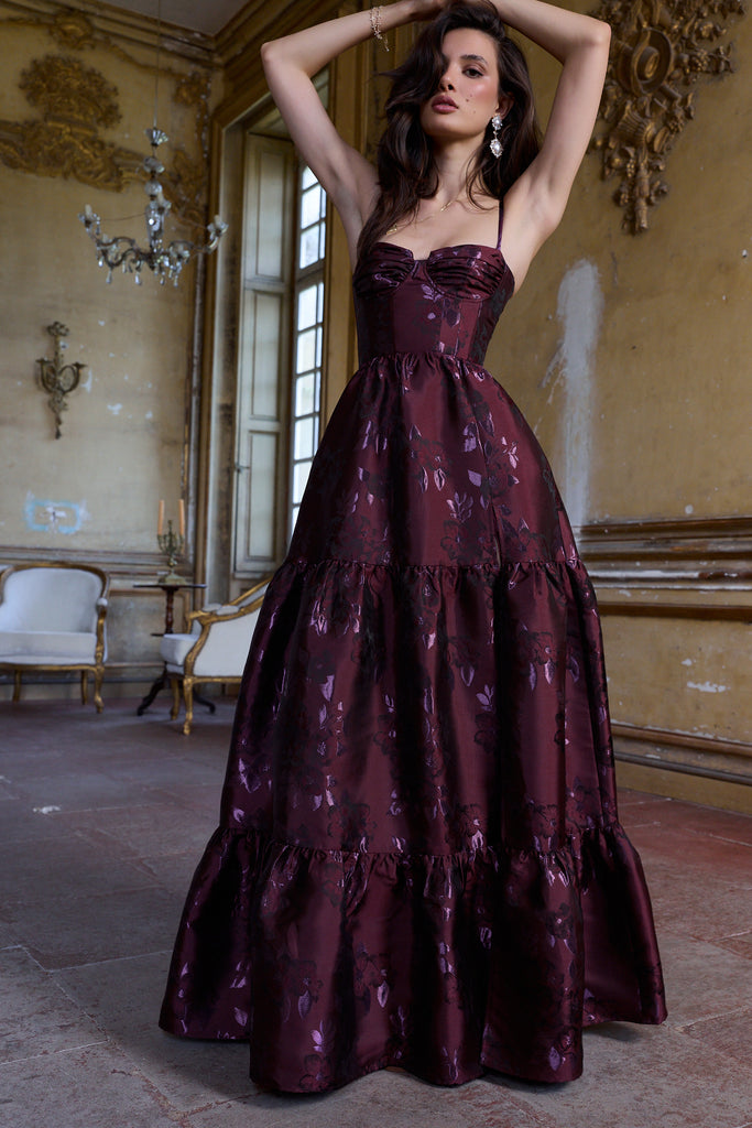 Woman in a burgundy evening gown standing in an elegant room with chandeliers.