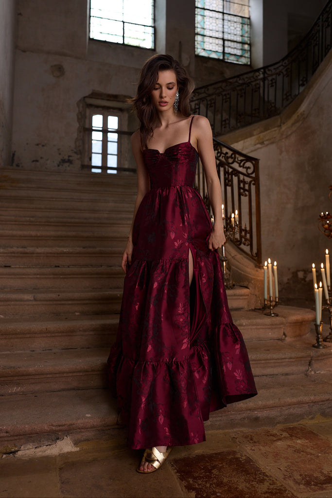 Woman in a burgundy evening gown standing on a staircase in an elegant interior setting.
