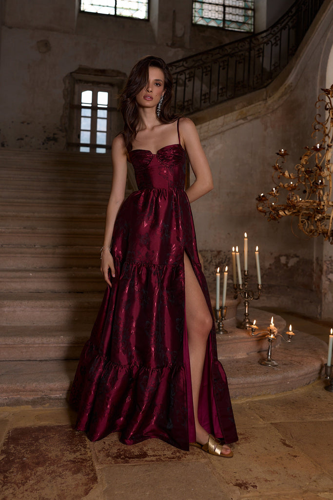 Woman in a burgundy evening gown standing in an elegant interior setting with candles and chandeliers.