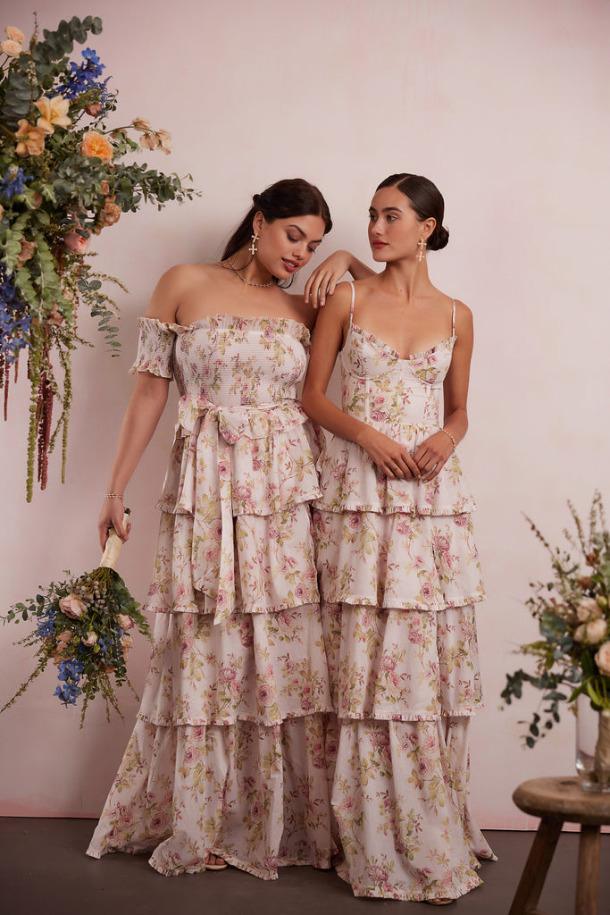 Two models wearing V.Chapman dresses in Pink Rose Print. Left model wearing The Angelina Dress showing full front view holding bouquet of flowers. Right model wearing The Caterina Dress in Pink Rose Print showing full front view.