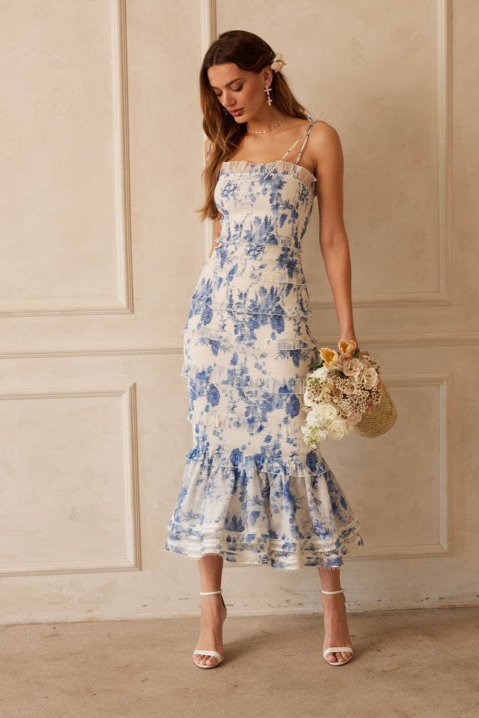 Walking Full front view of model wearing The Geranium Dress in Provencal Blue Floral while holding a basket of flowers.